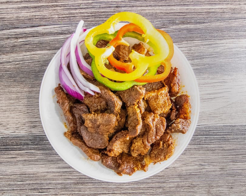 Plate of cooked meat pieces garnished with red onion slices and colorful bell peppers on a wooden table.