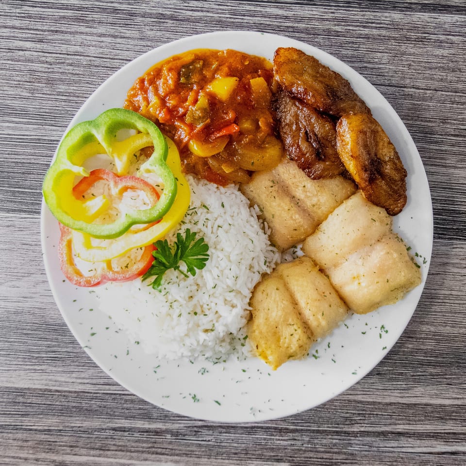 White plate with rice, fried fish fillets, plantains, vegetable stew, and colorful bell peppers.