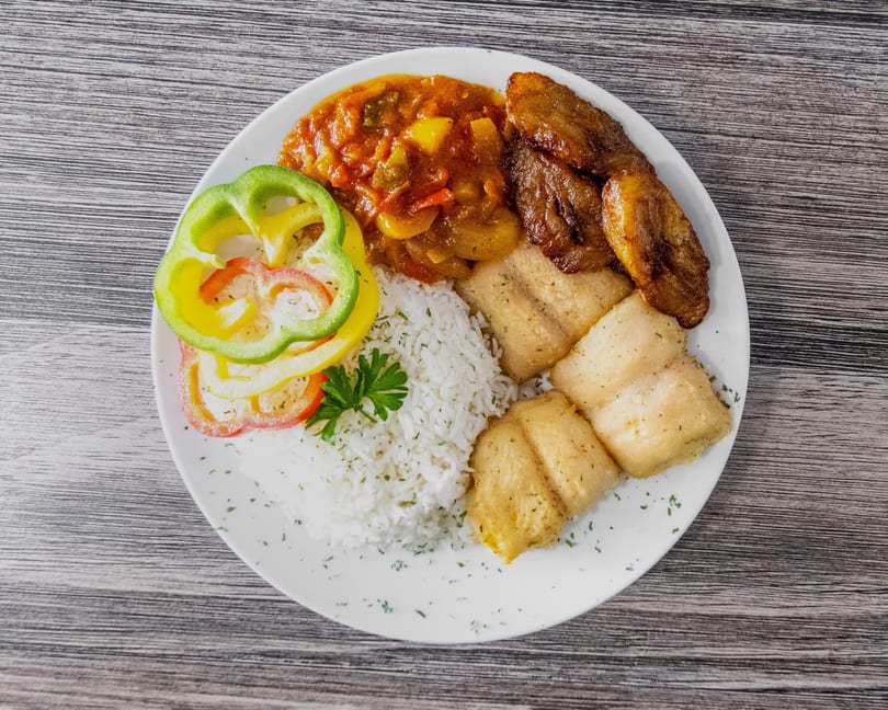 White plate with rice, fried fish fillets, plantains, vegetable stew, and colorful bell peppers.