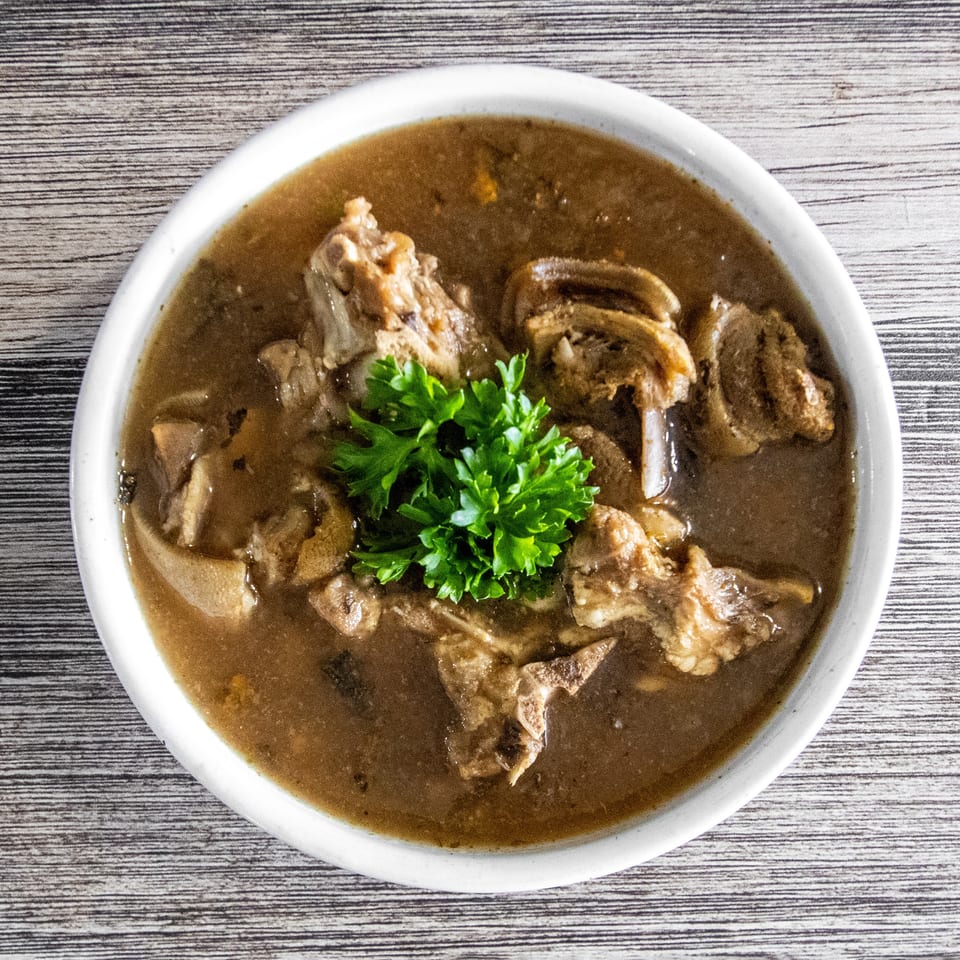 Overhead view of a rich brown stew with bone-in meat, tripe, and fresh parsley in a white bowl.