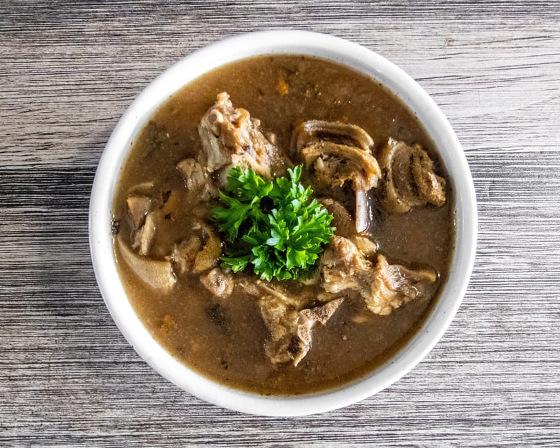 Overhead view of a rich brown stew with bone-in meat, tripe, and fresh parsley in a white bowl.