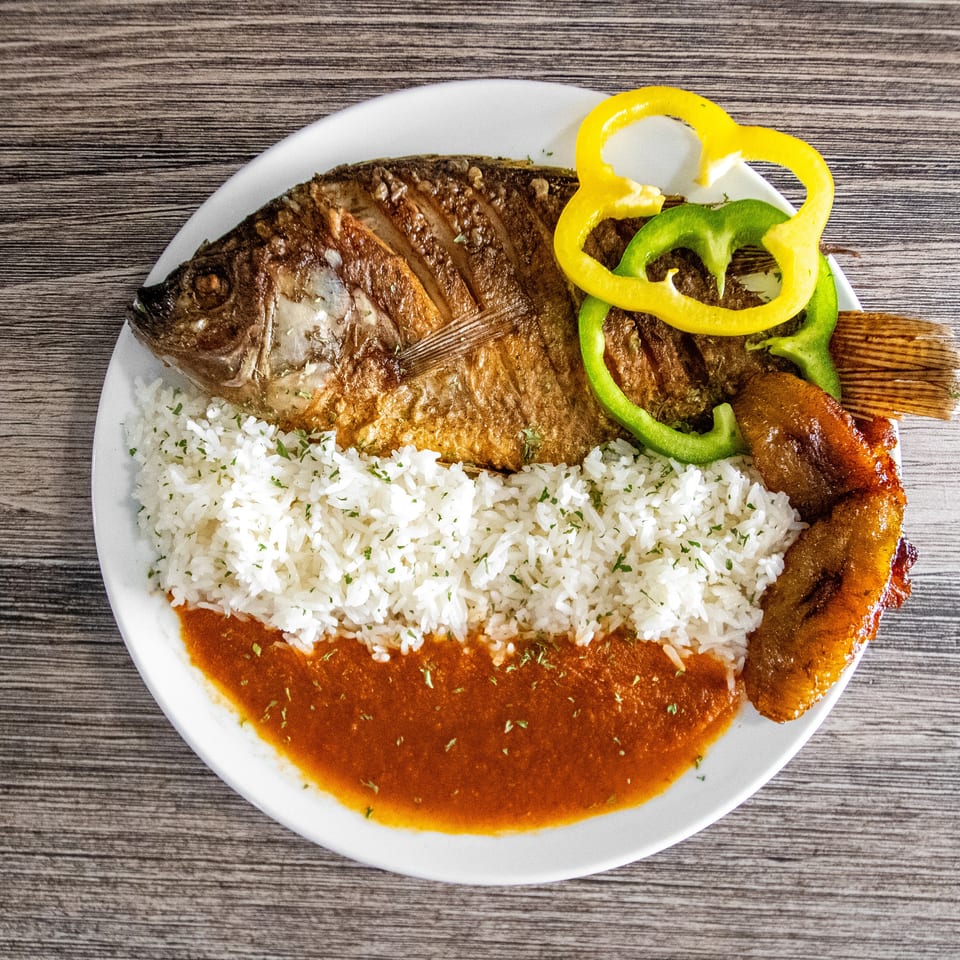 Whole fried fish with white rice, red sauce, bell peppers, and fried plantains on a white plate.
