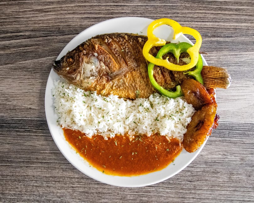 Whole fried fish with white rice, red sauce, bell peppers, and fried plantains on a white plate.