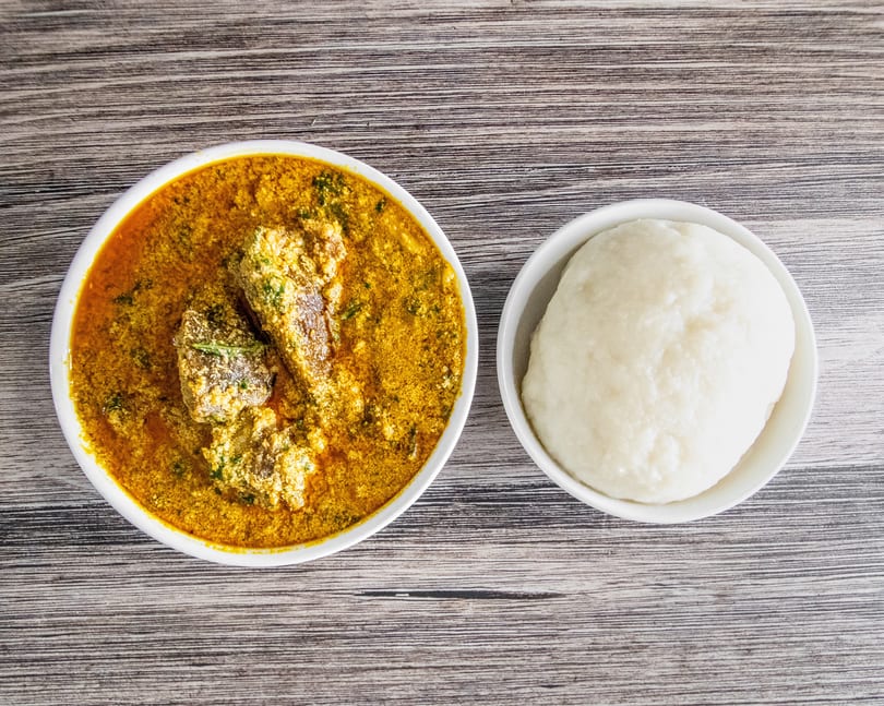 Overhead view of a bowl of Egusi soup with meat and a side of fufu on a wooden table.
