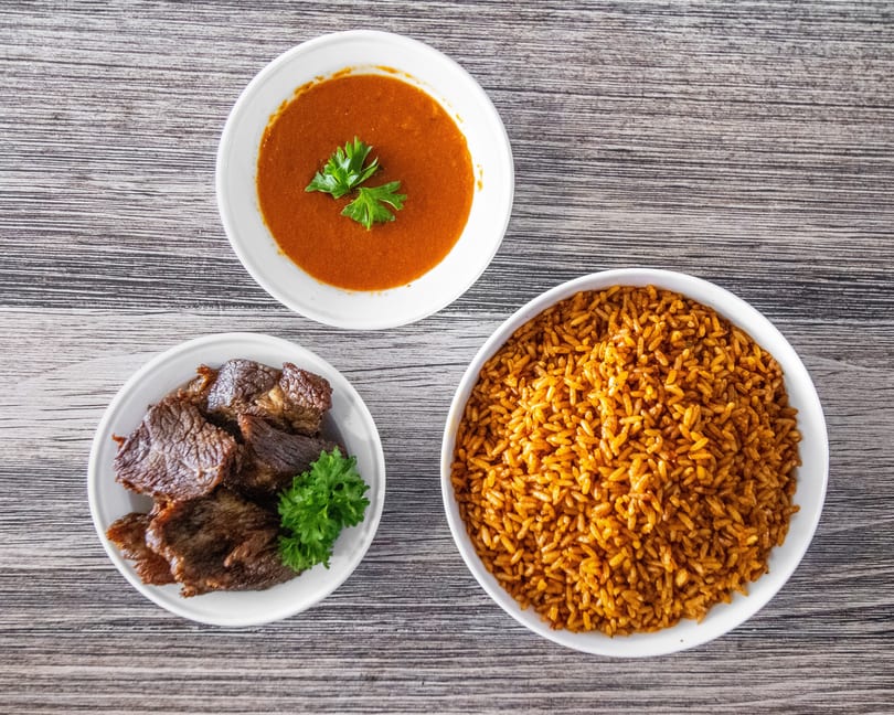 Three bowls of food: Jollof rice, stewed meat with parsley, and red soup on a wooden table.