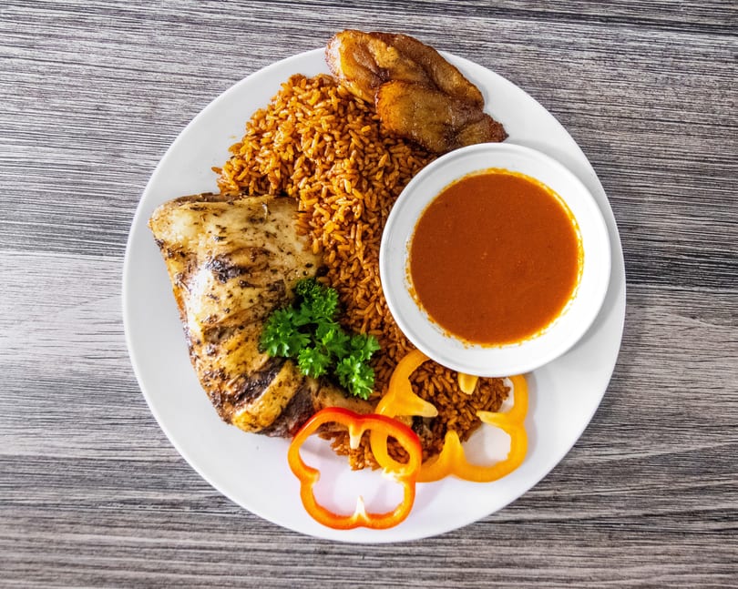 Grilled chicken, jollof rice, fried plantains, and red sauce on a white plate.