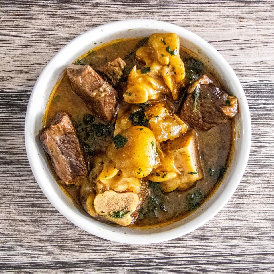 Hearty stew with beef chunks, cow foot, and greens in a white bowl on wood.