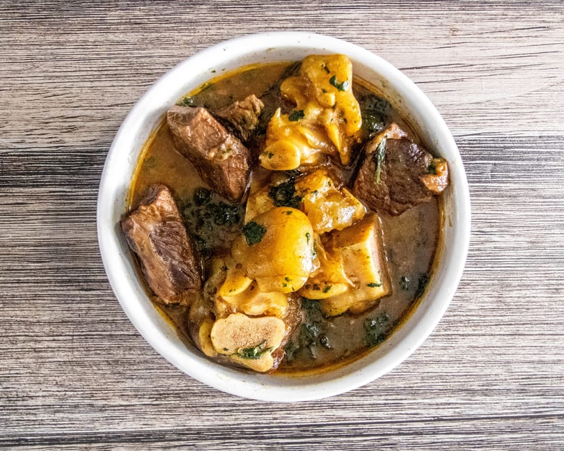Hearty stew with beef chunks, cow foot, and greens in a white bowl on wood.